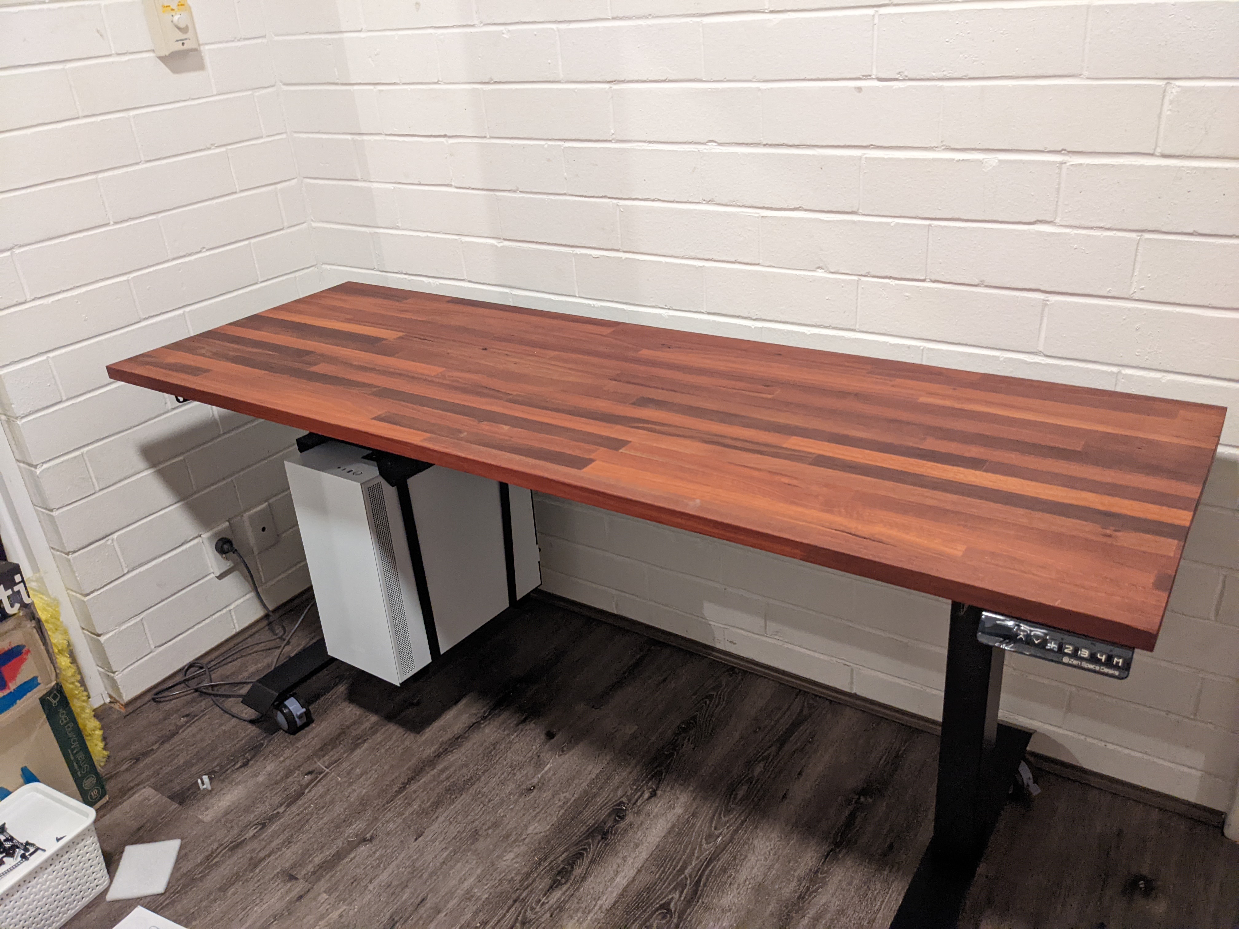 A fully oiled desk top attached to the top of a black metal desk frame with a white desktop computer mounted underneath it, there are controls on the right hand side of the desk to control the height
