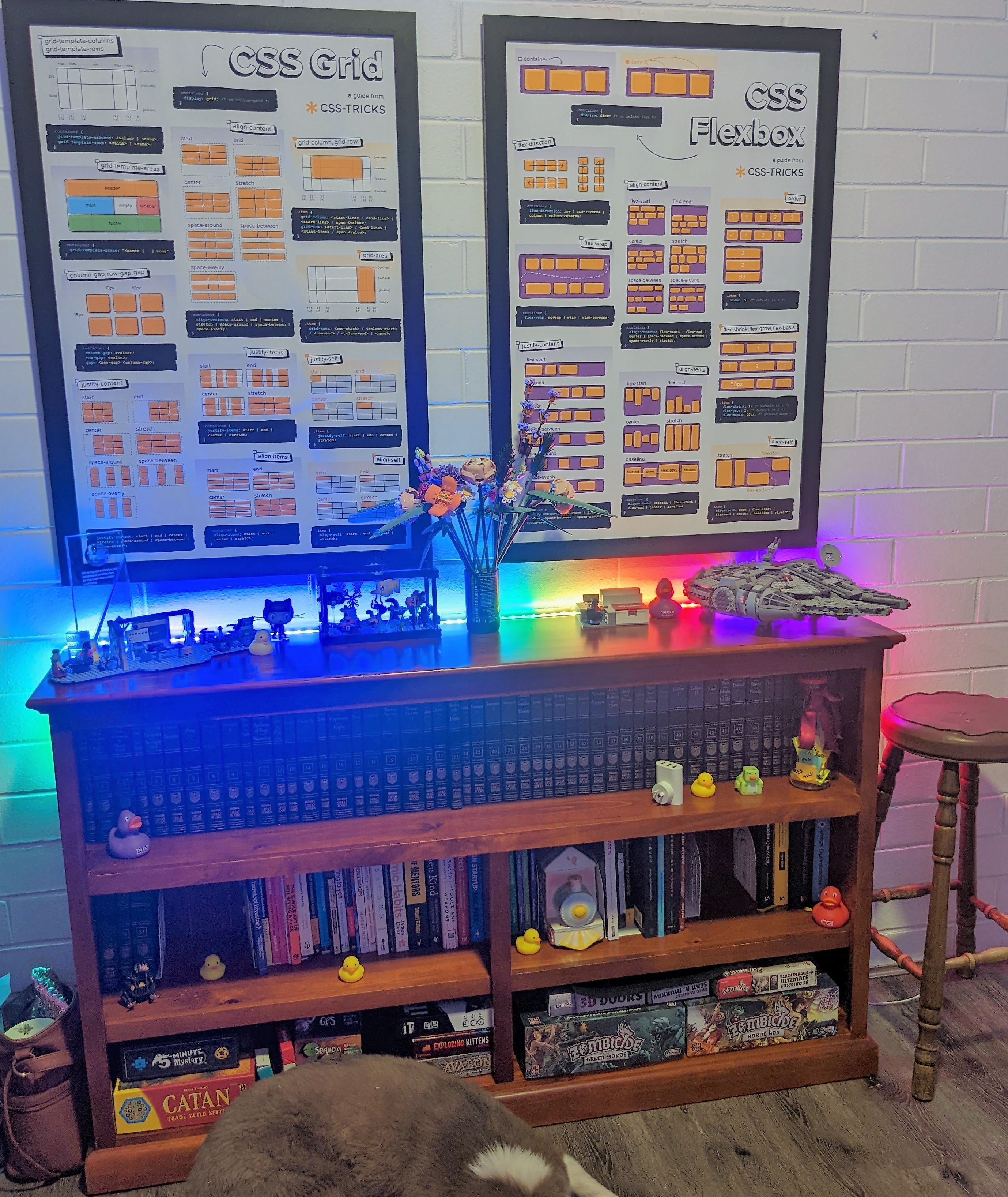 Two posters with information and graphics about CSS Grid and Flexbox, positioned above a bookshelf full of books and boardgames, with various assembled Lego sets on top of the shelf and rubber ducks positioned around the shelf